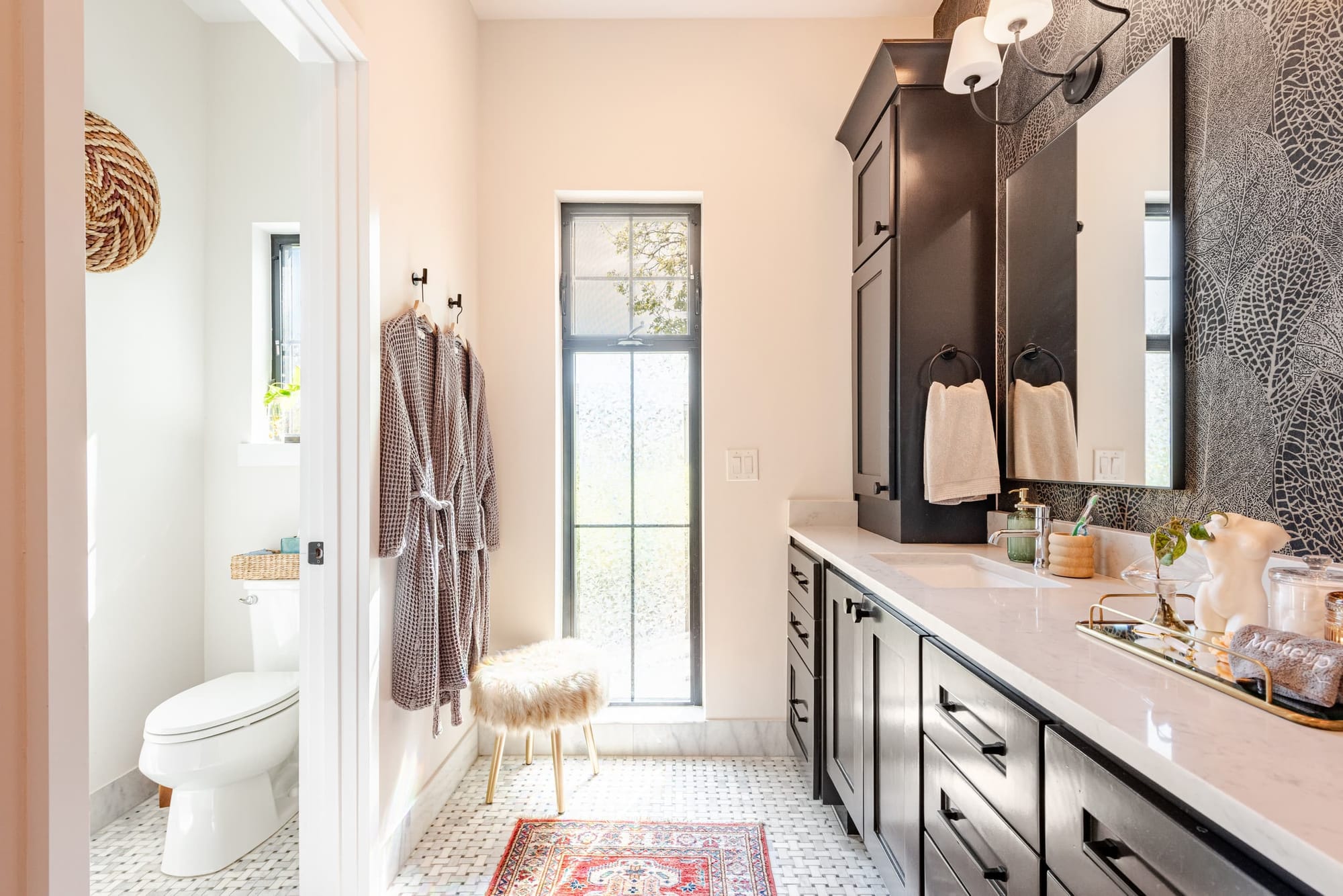 A bath with a black double vanity and marble counters, waffle robes on hooks, and a vintage red runner on penny tile