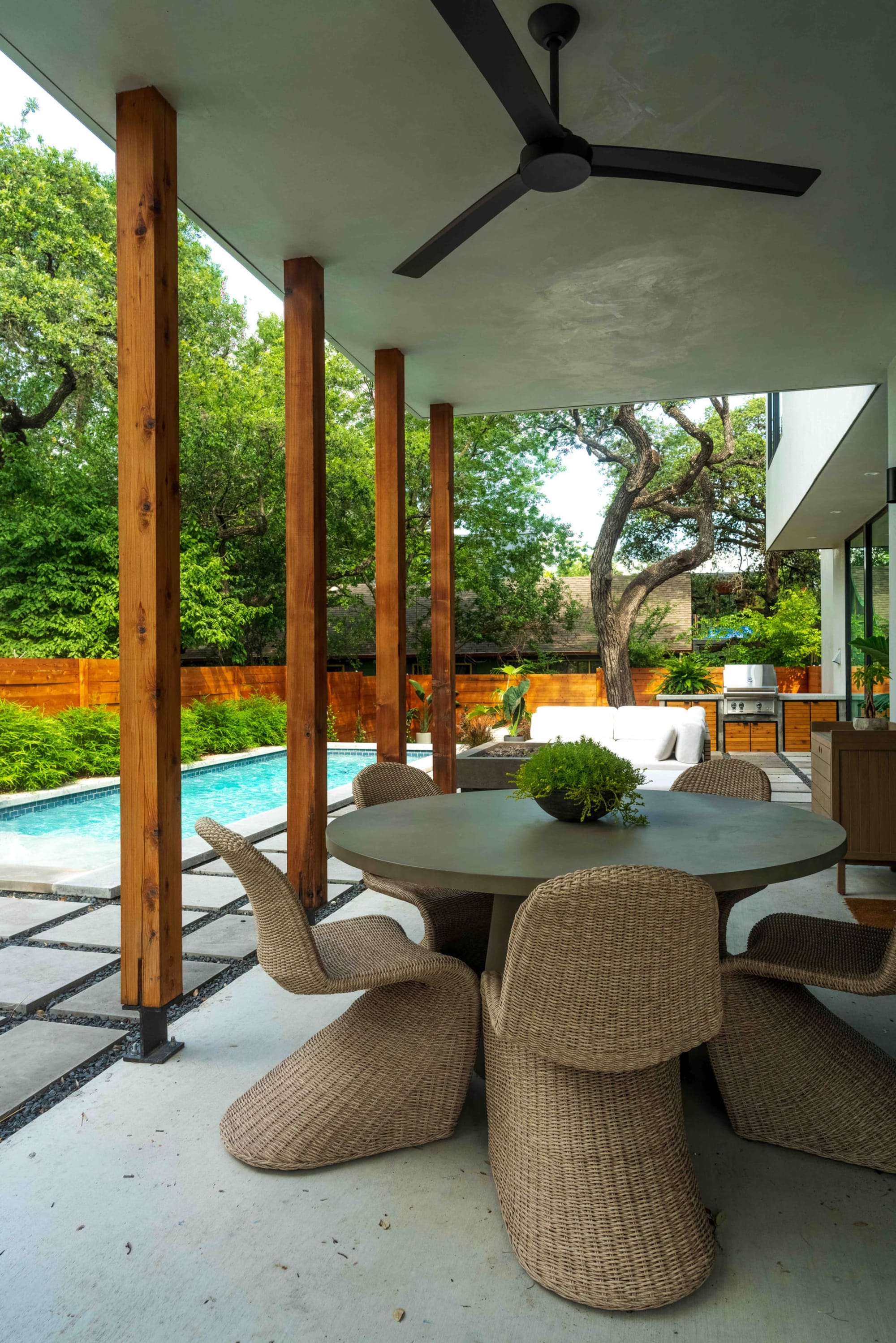 Covered outdoor dining — a round stone-top table and woven chairs under cedar posts, beside the pool and outdoor kitchen