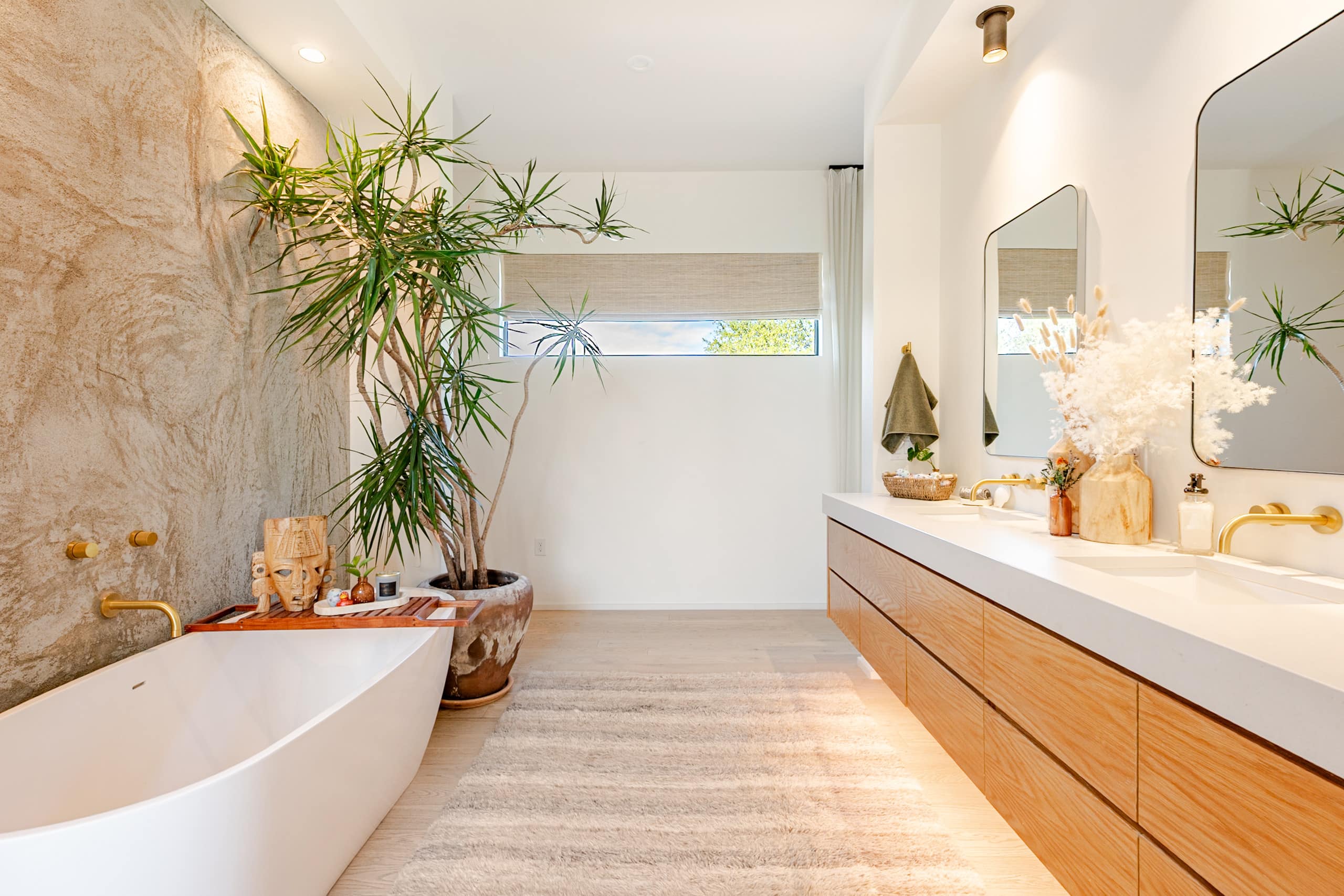 The primary bath — a freestanding soaking tub against a limewash plaster wall, a floating oak double vanity, and a potted dracaena