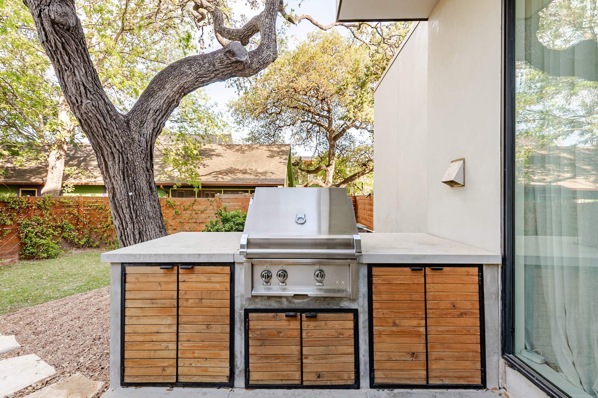 The custom built-in outdoor grill station, concrete-topped with wood-front cabinets, beneath a sprawling live oak