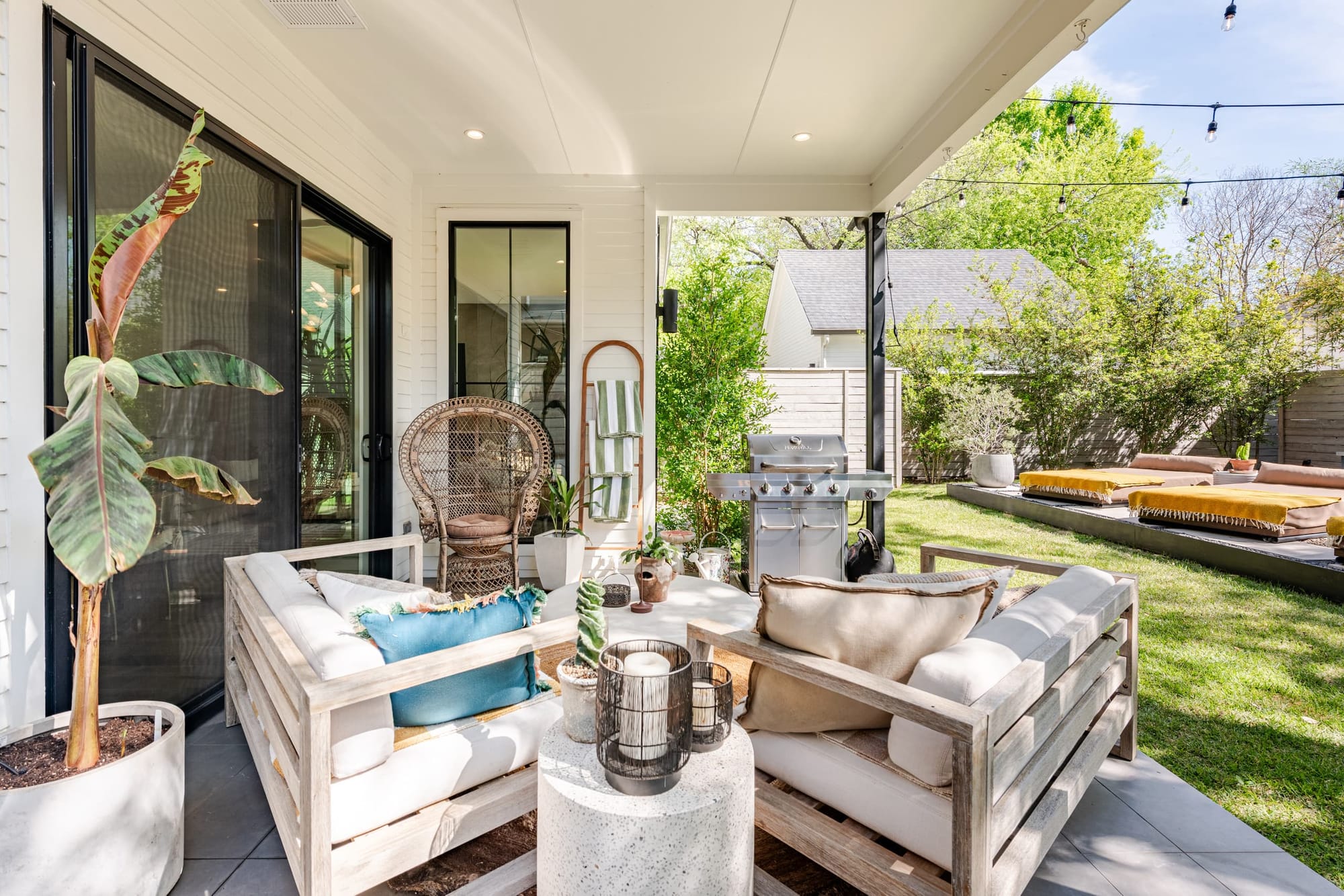 The covered back porch — whitewashed timber sofas, a peacock chair, and string lights over the lawn