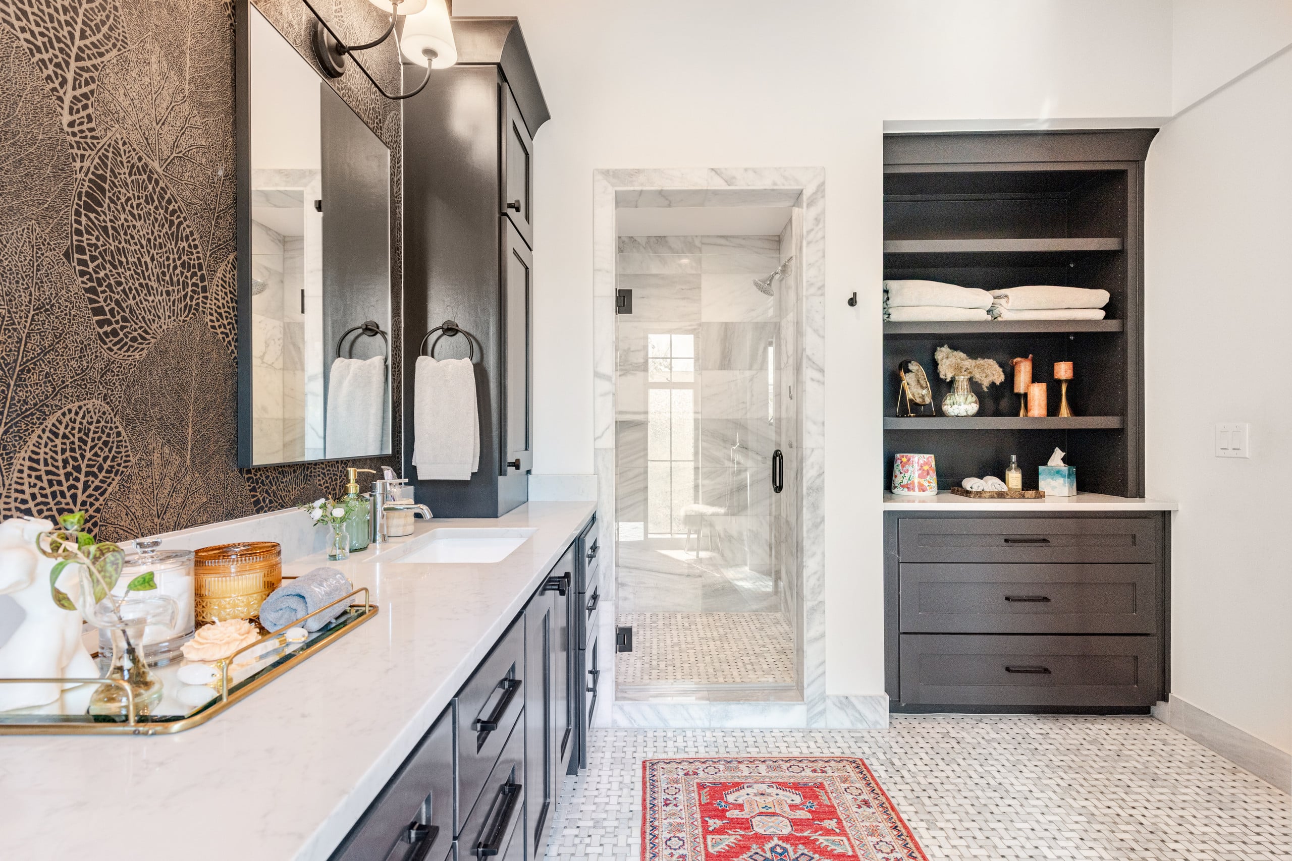 The primary bath at The Hether Haus — a black double vanity with marble counters, a built-in linen cabinet, and the marble walk-in shower beyond